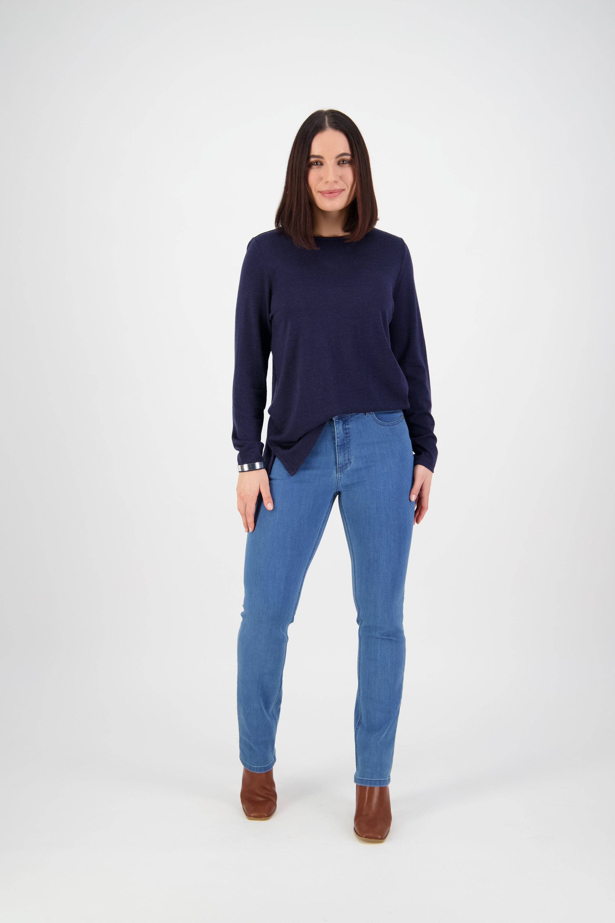 A woman with straight dark hair stands facing forward against a plain white background, wearing a navy sweater, Vassalli Onset Jean - Plain New Blue, and brown ankle boots.