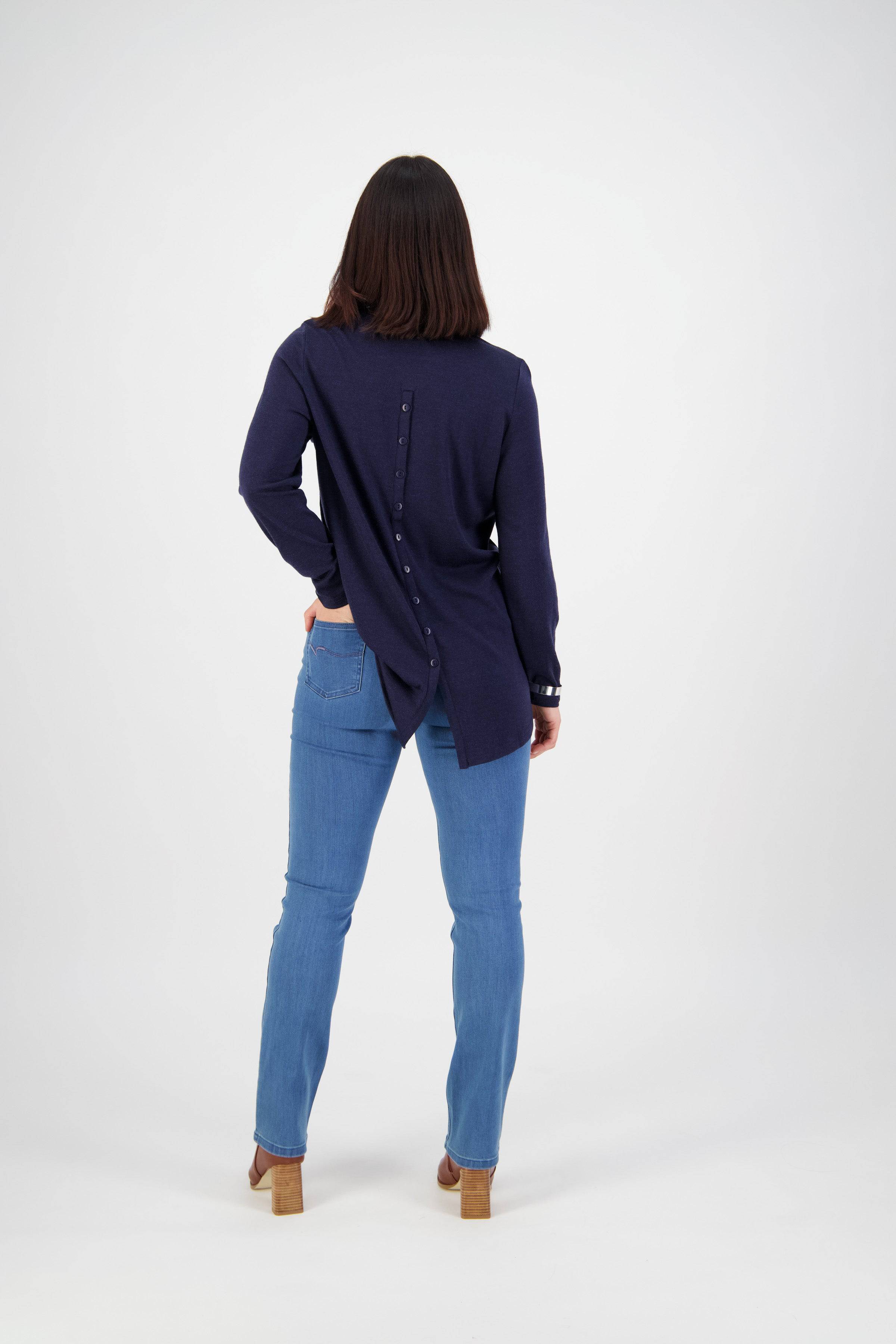 A person with shoulder-length dark hair faces away, wearing a long-sleeve navy top with back buttons and Vassalli’s Onset Jean - Plain New Blue. They wear light brown heeled shoes and stand against a plain white background.