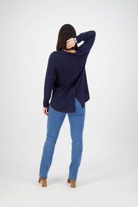 A woman faces away, wearing a navy long-sleeve top with back buttons, Vassalli Onset Jean - Plain New Blue, and tan heels. She rests one hand on the back of her head and has straight dark hair.