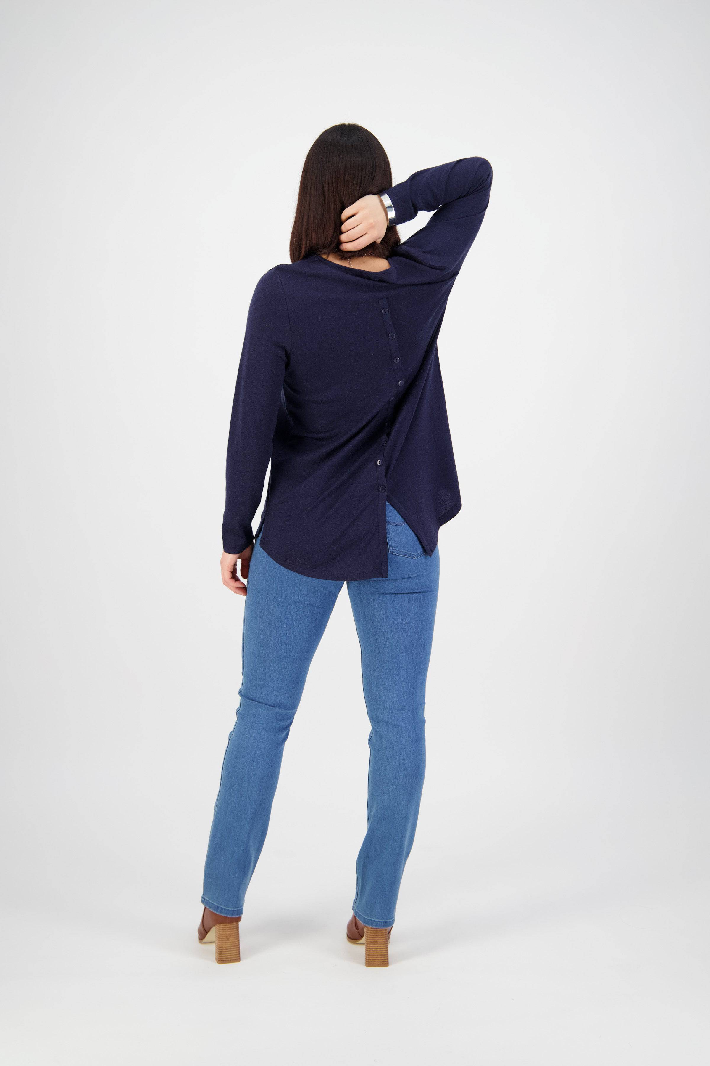 A woman faces away, wearing a navy long-sleeve top with back buttons, Vassalli Onset Jean - Plain New Blue, and tan heels. She rests one hand on the back of her head and has straight dark hair.