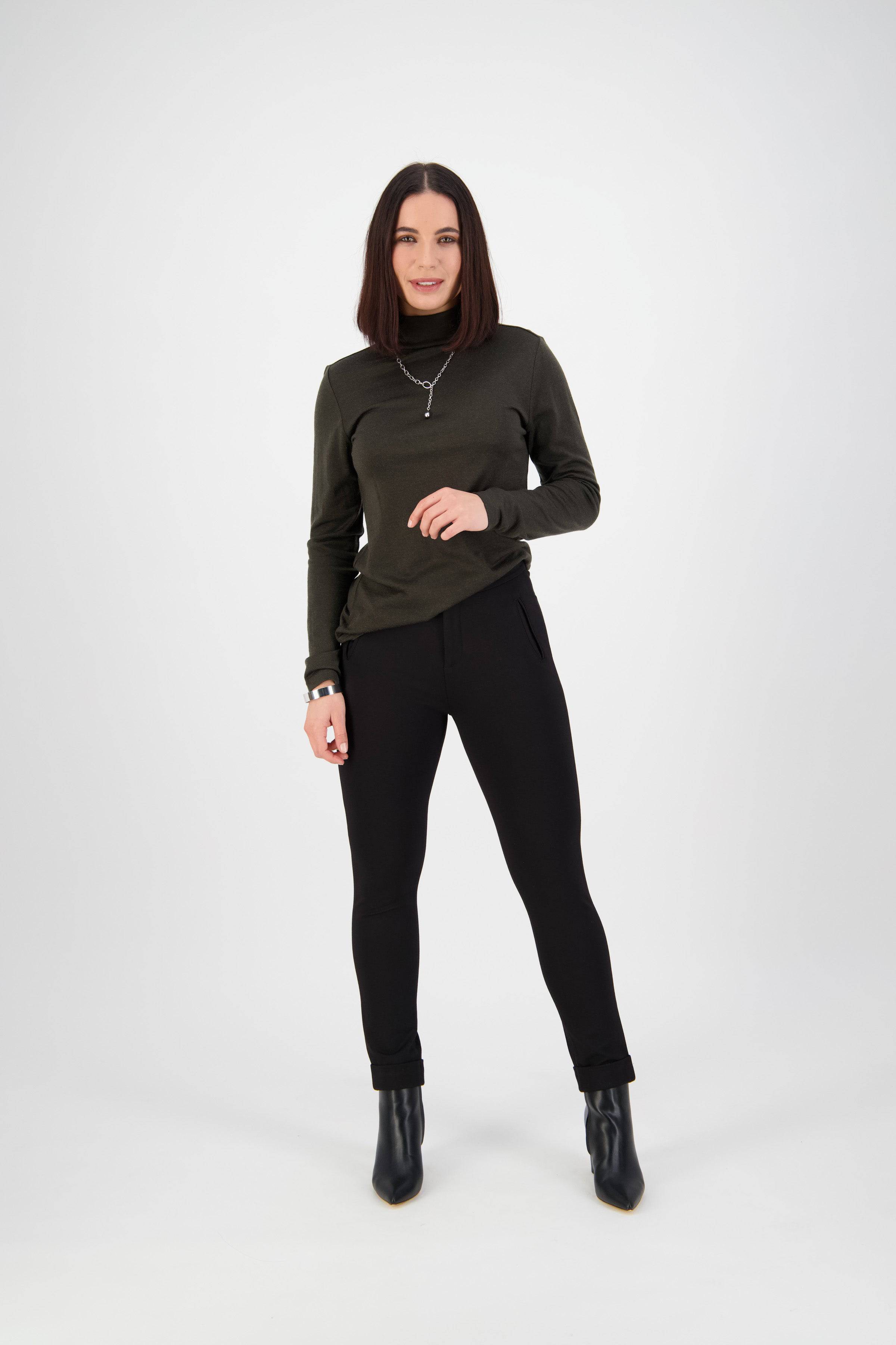A woman with straight, shoulder-length brown hair stands in a studio against a white background, wearing a dark green long-sleeve turtleneck, Vassalli’s Style Pant - Black, black ankle boots, and a necklace, smiling slightly.