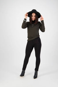 A woman stands against a plain white background, smiling while holding her black wide-brim hat. She wears a dark long-sleeve top and Vassalli Style Pant - Black, paired with black ankle boots.