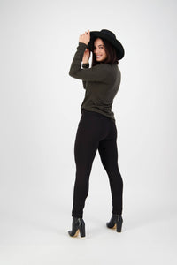 A smiling woman in a black wide-brim hat, dark green long-sleeve top, and Vassalli Style Pant - Black poses in a studio, holding her hat and looking over her shoulder.