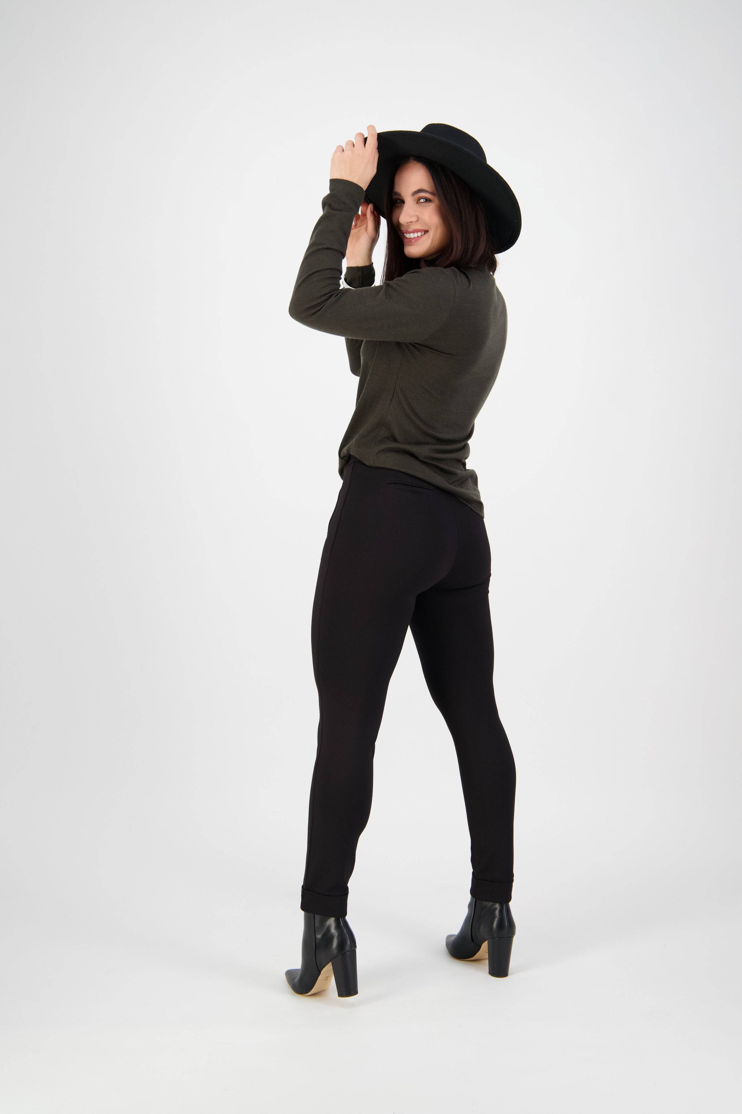 A smiling woman in a black wide-brim hat, dark green long-sleeve top, and Vassalli Style Pant - Black poses in a studio, holding her hat and looking over her shoulder.
