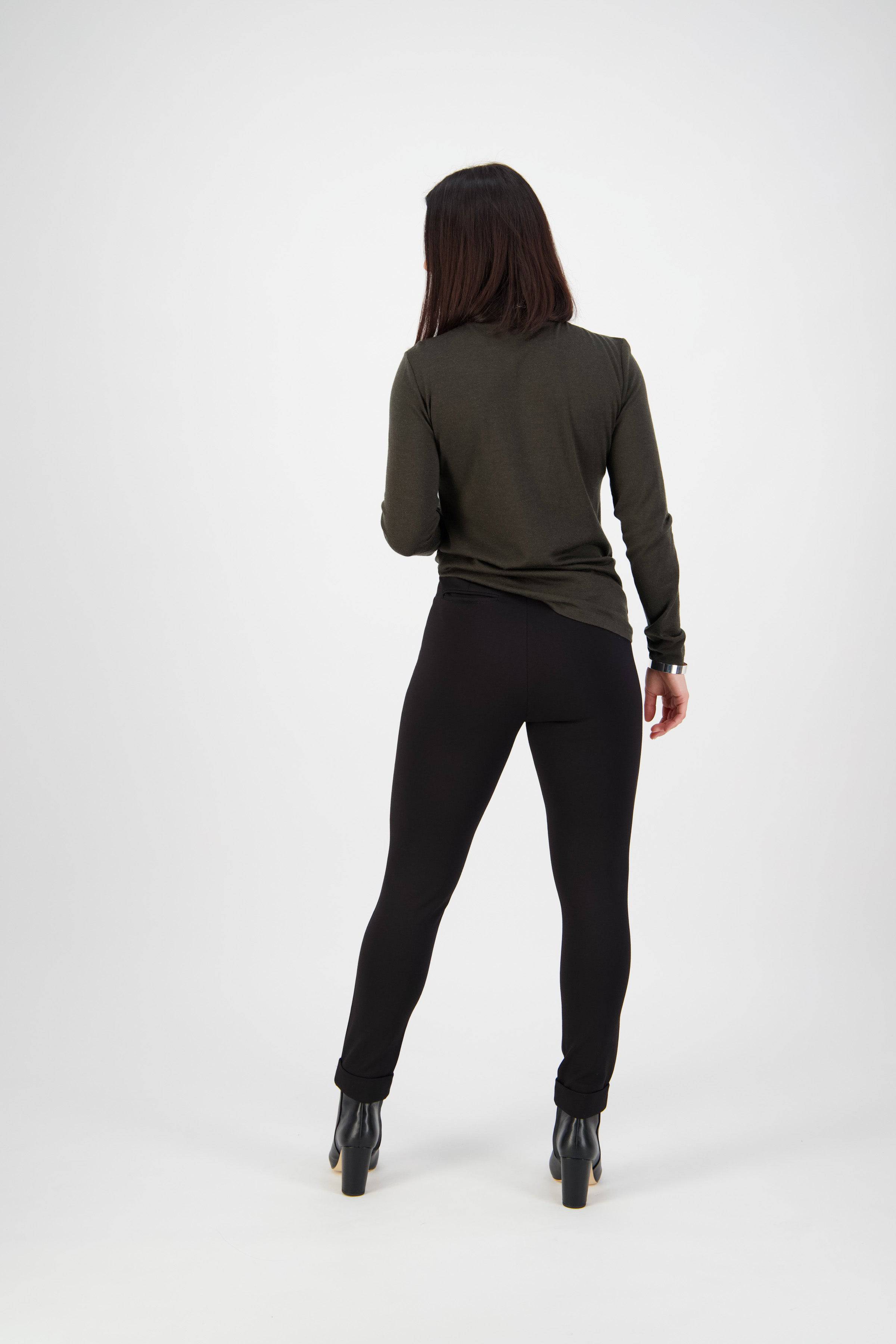 A woman with straight dark hair faces away, wearing a long-sleeved dark green top and Vassalli Style Pant - Black, made from stretch fabric, paired with black heeled boots against a plain light background.