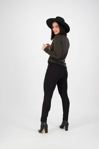 A woman in a black wide-brim hat, olive green sweater, and Vassalli Style Pant - Black smiles and looks back over her shoulder against a plain white background.