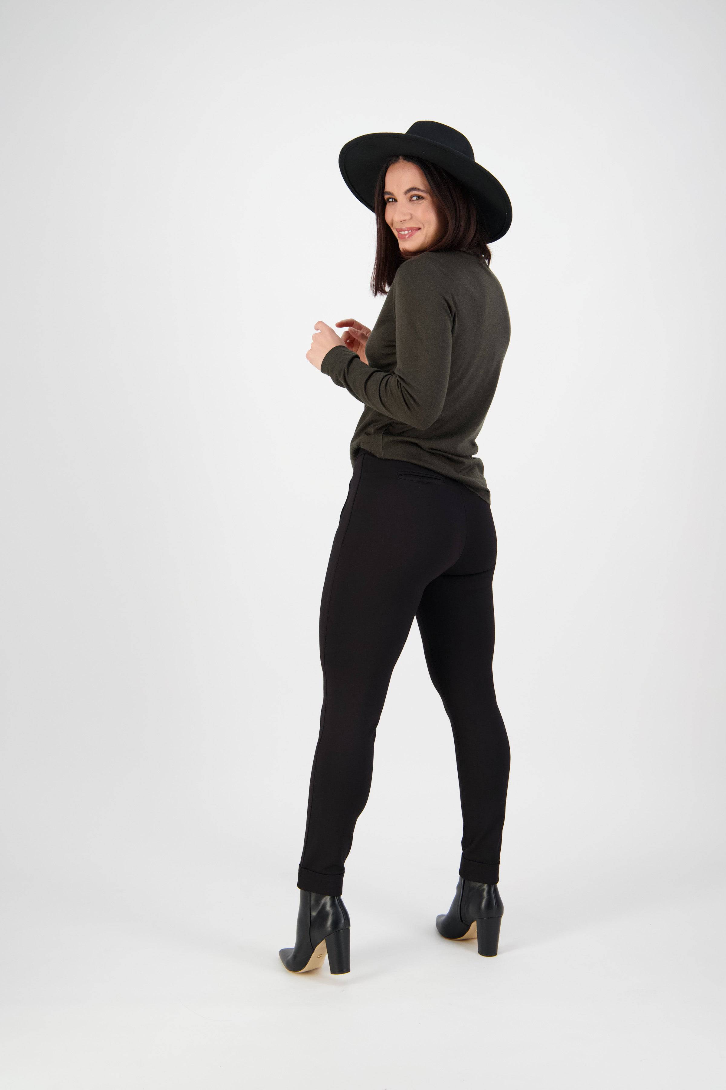 A woman in a black wide-brim hat, olive green sweater, and Vassalli Style Pant - Black smiles and looks back over her shoulder against a plain white background.