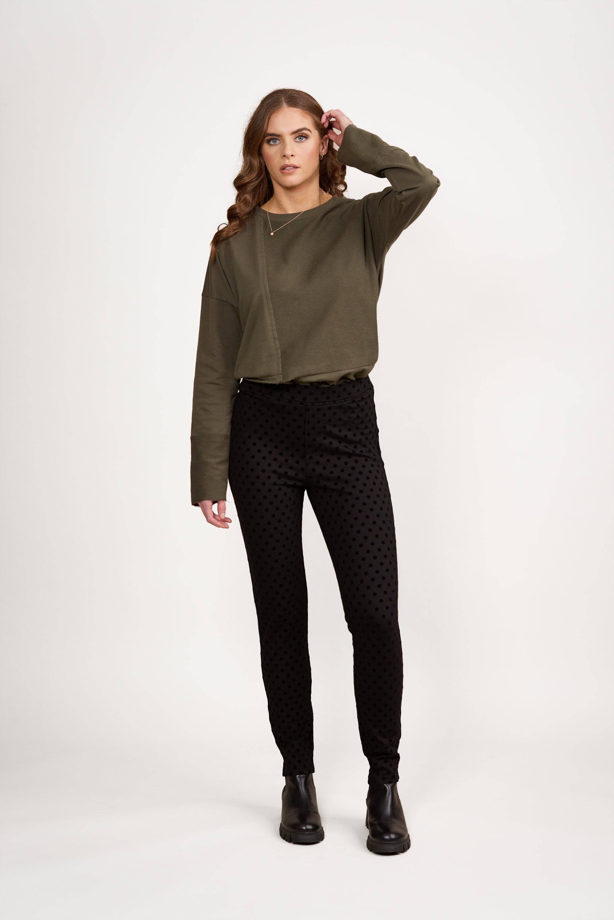 A woman with long brown hair stands against a white background, wearing an olive green long-sleeve top and Vassalli's View Pant - Black, a skinny leg ankle grazer. She has a neutral expression, her left hand touching her head.