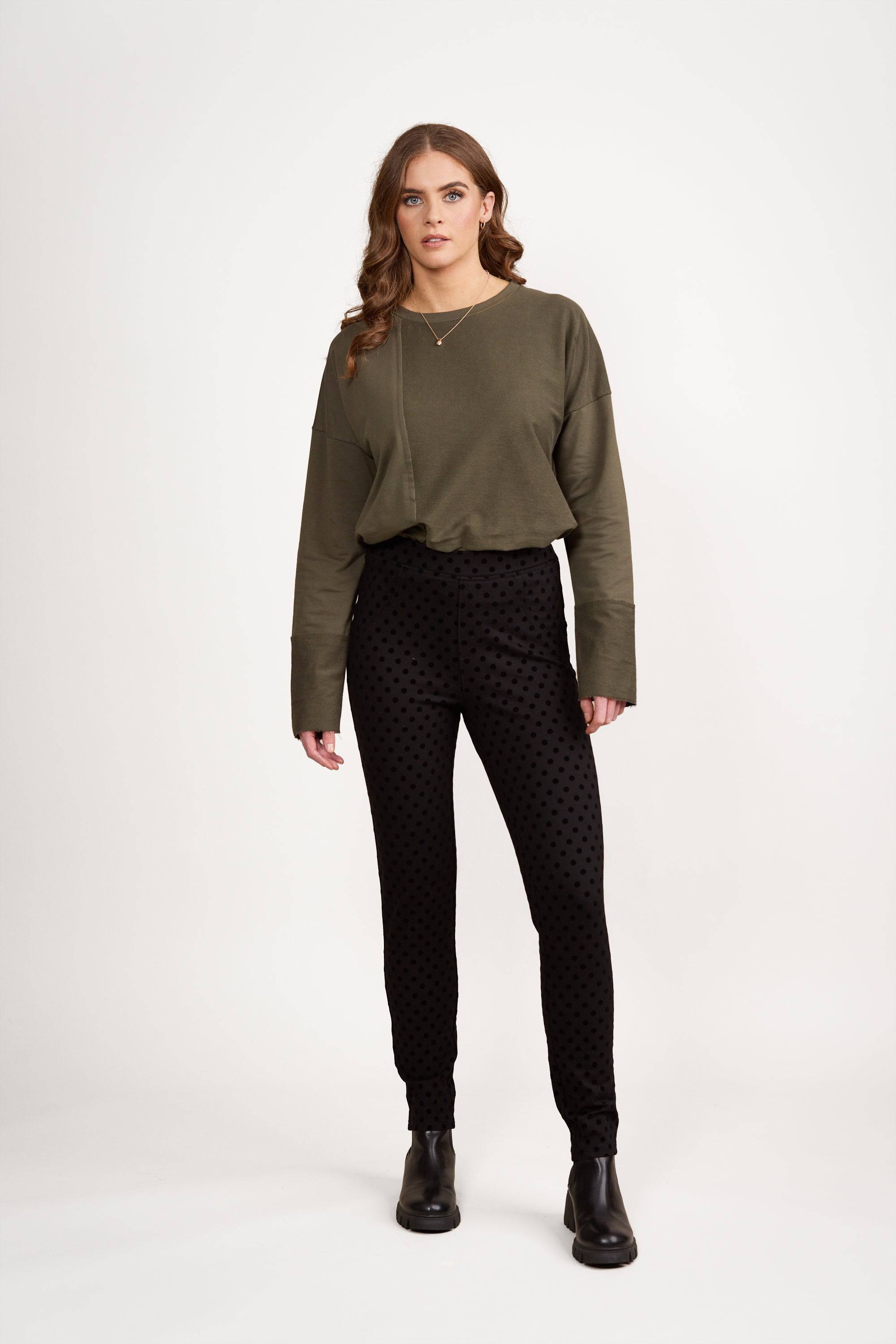 A woman with long wavy hair stands against a white background wearing an olive green long-sleeve top, Vassalli View Pant - Black with a skinny leg ankle grazer fit, and black ankle boots. She has a neutral expression.