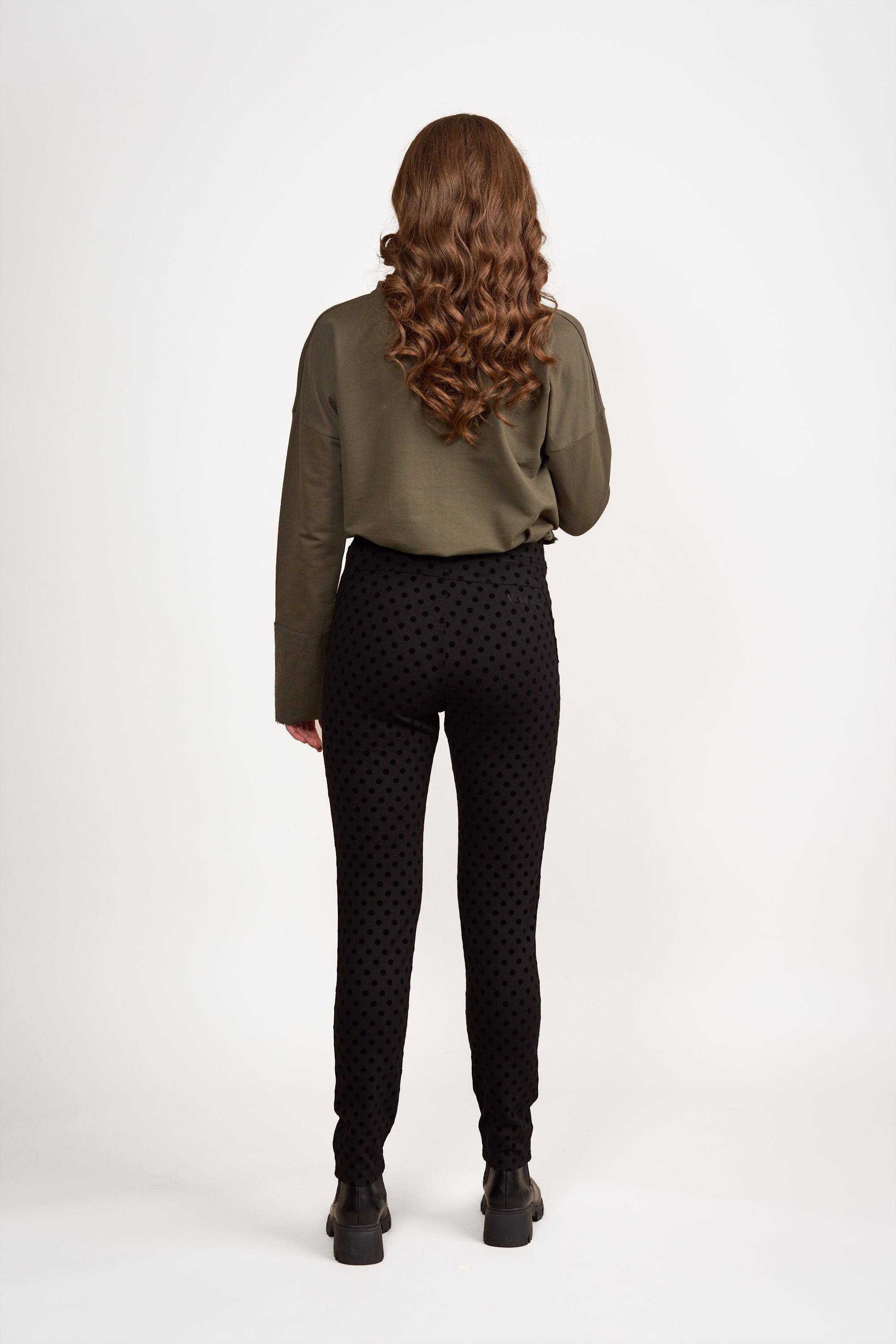 A person with long, wavy brown hair stands facing away, wearing an olive green long-sleeve top and Vassalli View Pant - Black, made from stretch fabric, paired with black shoes against a plain white background.