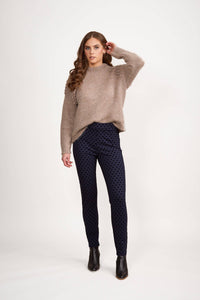 A woman with long brown hair wears a loose beige sweater, Vassalli View Pant in Navy, and black ankle boots, standing against a plain white background. She has one hand near her ear and looks at the camera.