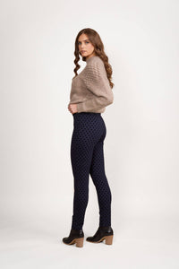 A woman with long, wavy brown hair stands sideways against a plain white background. She wears a beige sweater and Vassalli View Pant in Navy, styled as skinny leg ankle grazers, paired with black heeled ankle boots, looking over her shoulder.