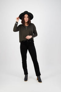 A woman poses against a white background, wearing a black wide-brim hat, a dark green long-sleeve top, and Vassalli's Arch Cord Pant in black with a straight leg and stretch fabric, looking at the camera while holding her hat brim.