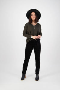 A person stands against a plain white background, wearing a wide-brimmed black hat, olive green long-sleeve shirt, Vassalli Arch Cord Pant in black, and black ankle boots. They have shoulder-length dark hair and a neutral expression.
