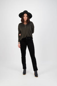 A woman, smiling with her arms behind her back, wears a black wide-brimmed hat, dark long-sleeve top, black boots, and Vassalli Arch Cord Pant in black. She stands against a plain white background.