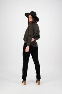 A woman in a black wide-brim hat, dark long-sleeve top, and Vassalli’s Arch Cord Pant - Black stands facing away while looking back over her shoulder, posing against a plain white background.