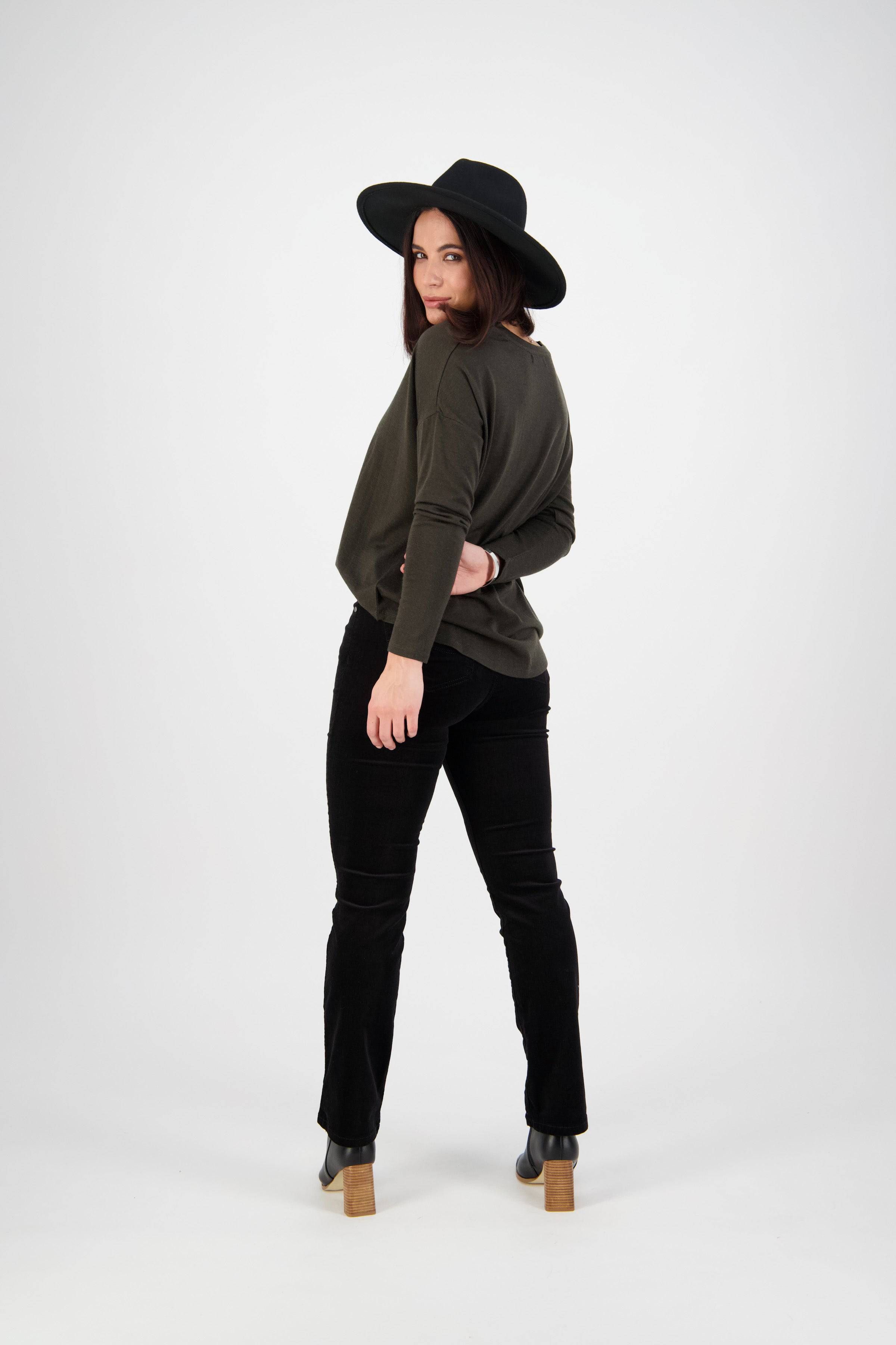 A woman in a black wide-brim hat, dark long-sleeve top, and Vassalli’s Arch Cord Pant - Black stands facing away while looking back over her shoulder, posing against a plain white background.