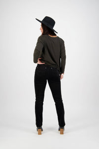 A person stands facing away from the camera, wearing a black wide-brimmed hat, dark long-sleeve shirt, Vassalli Arch Cord Pant - Black with a straight leg and stretch fabric, and wooden-heeled shoes against a plain white background.