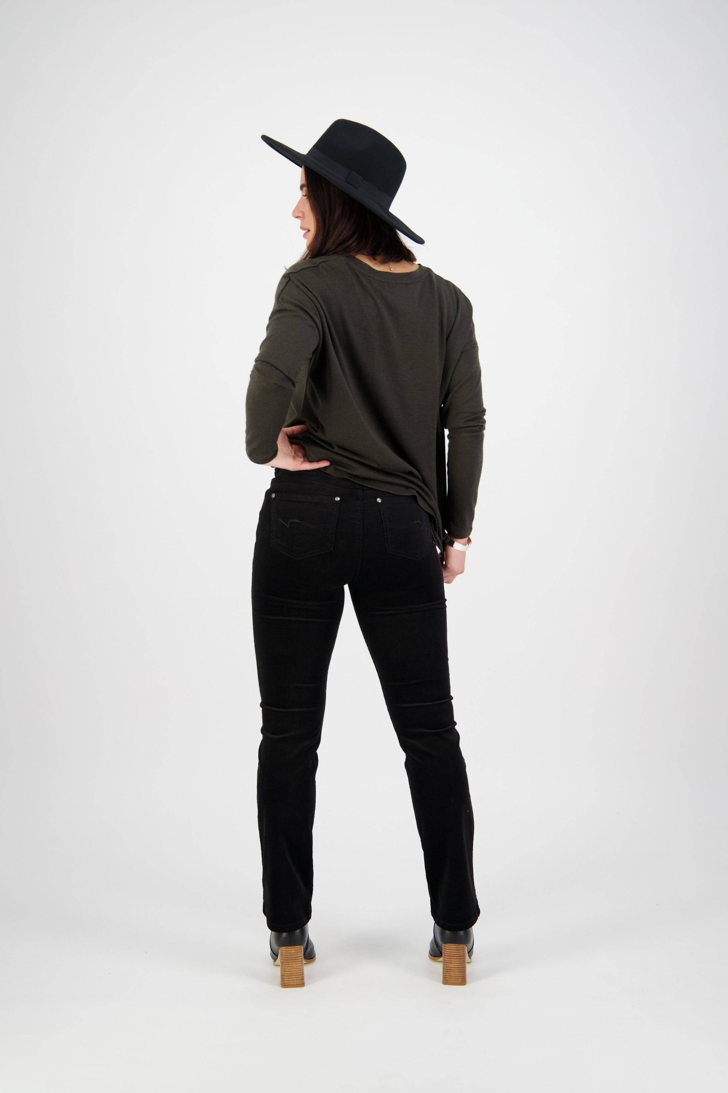 A person stands facing away from the camera, wearing a black wide-brimmed hat, dark long-sleeve shirt, Vassalli Arch Cord Pant - Black with a straight leg and stretch fabric, and wooden-heeled shoes against a plain white background.