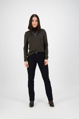 A woman with straight dark hair faces forward in a dark green turtleneck, Vassalli Tour Jean - Black Denim, black boots, and a silver necklace against a plain white background.
