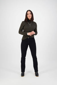 A woman with straight, shoulder-length dark hair stands against a white background, wearing a dark green long-sleeve top, Vassalli Tour Jean in Black Denim, black ankle boots, and a silver necklace. She smiles softly with her hands clasped.