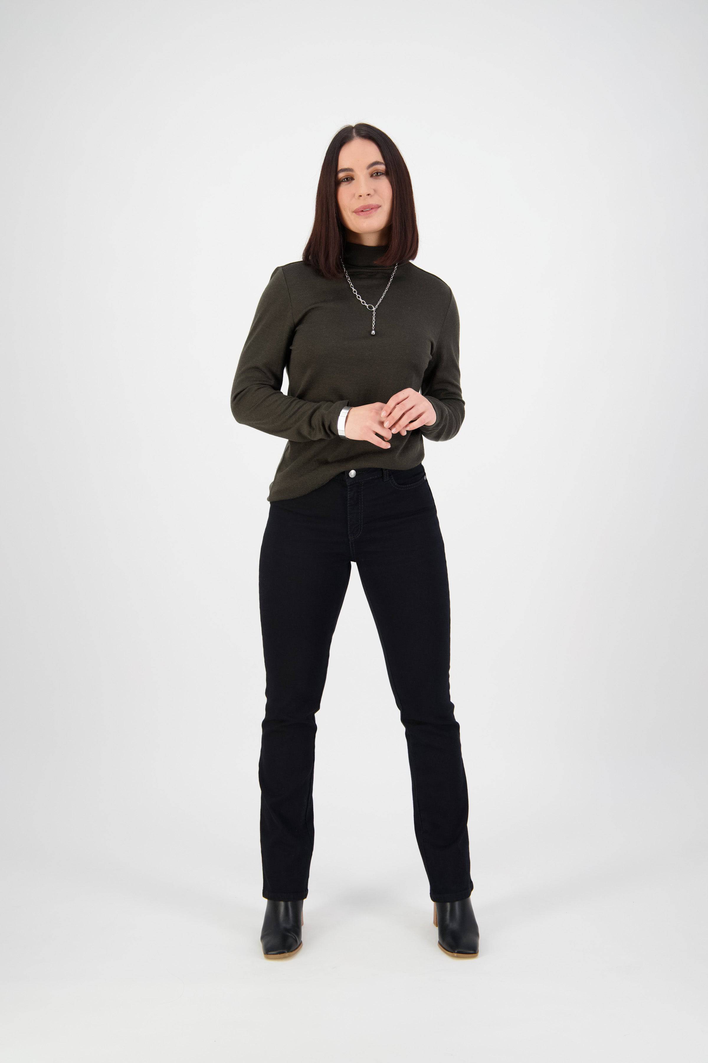 A woman with straight, shoulder-length dark hair stands against a white background, wearing a dark green long-sleeve top, Vassalli Tour Jean in Black Denim, black ankle boots, and a silver necklace. She smiles softly with her hands clasped.