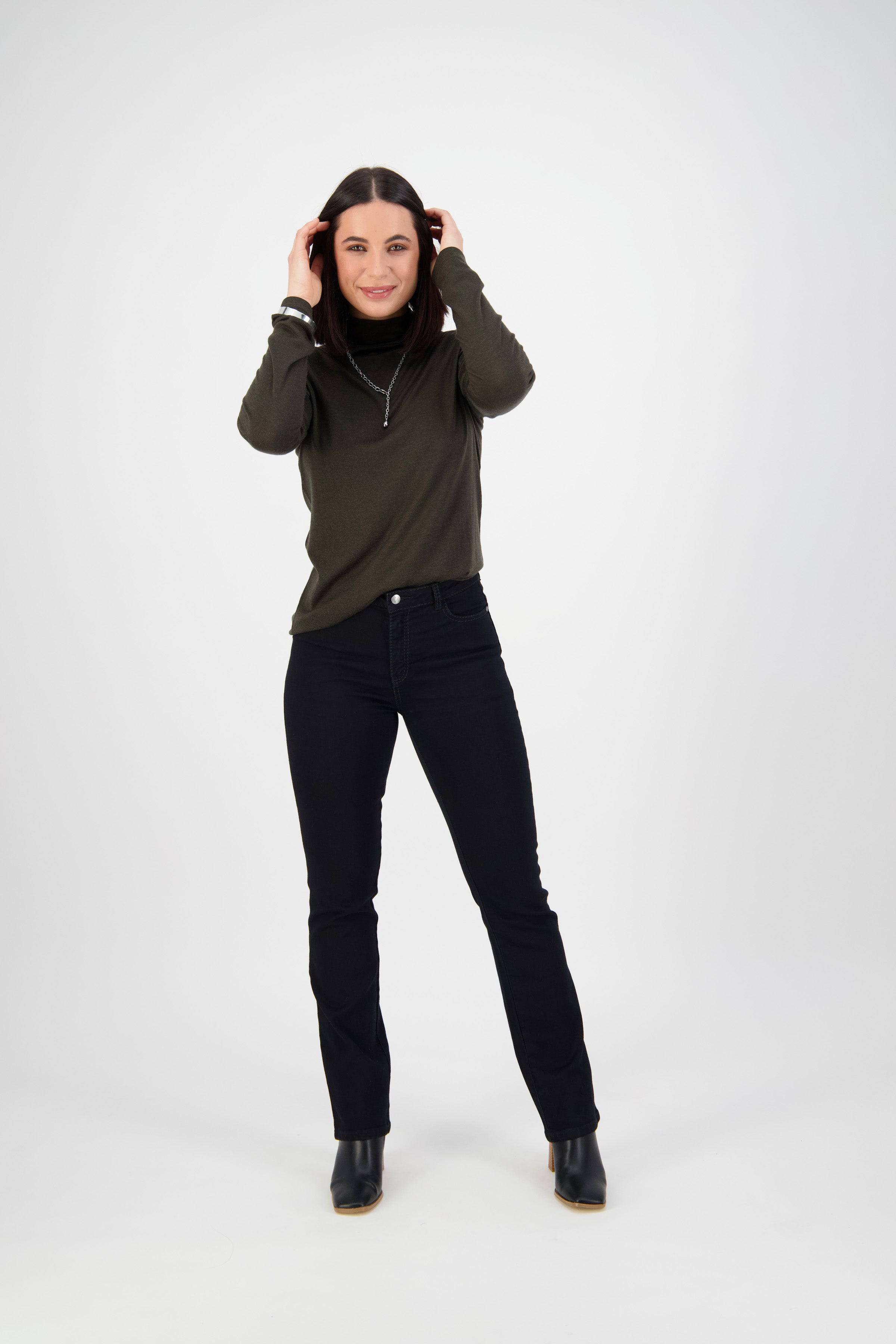 A woman with dark hair smiles in front of a white background, wearing a dark long-sleeve top, black boots, and Vassalli Tour Jean - Black Denim. She raises her hands to her head, touching her hair.