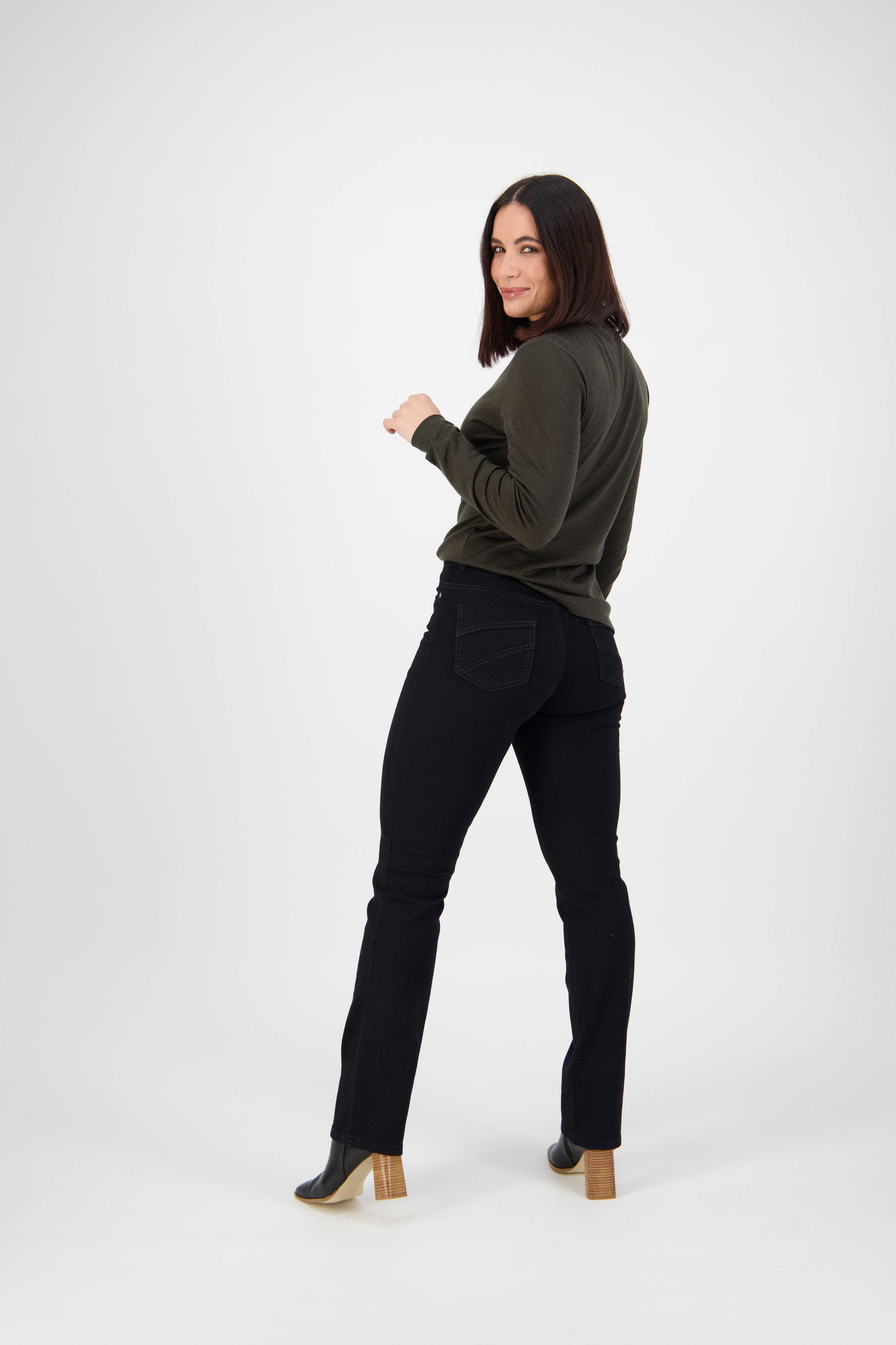 A woman with shoulder-length dark hair in a dark green long-sleeve top, Vassalli Tour Jean - Black Denim, and black heeled boots stands turning slightly left and smiling against a plain white background.