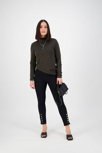 A woman with straight dark hair stands against a white background, wearing a dark green long-sleeve top, Vassalli Ankle Grazer Jean - Black Denim, black heeled sandals, and holding a black handbag.