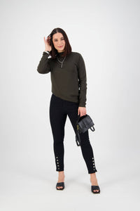 A woman with straight brown hair wears a dark green long-sleeve top, Vassalli Ankle Grazer Jean - Black Denim, and black open-toe shoes. She holds a small black handbag and smiles against a plain white background.