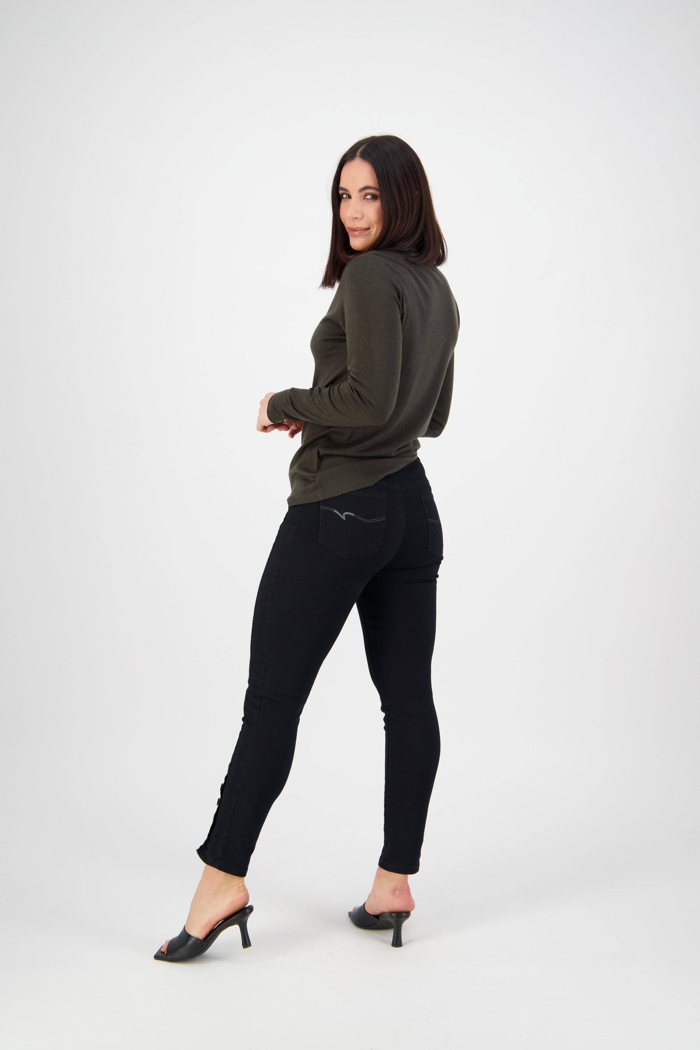 A woman with straight dark hair, in a dark green long-sleeve top, Vassalli Ankle Grazer Jean - Black Denim, and black heeled sandals stands facing away but looks back and smiles in a white studio.