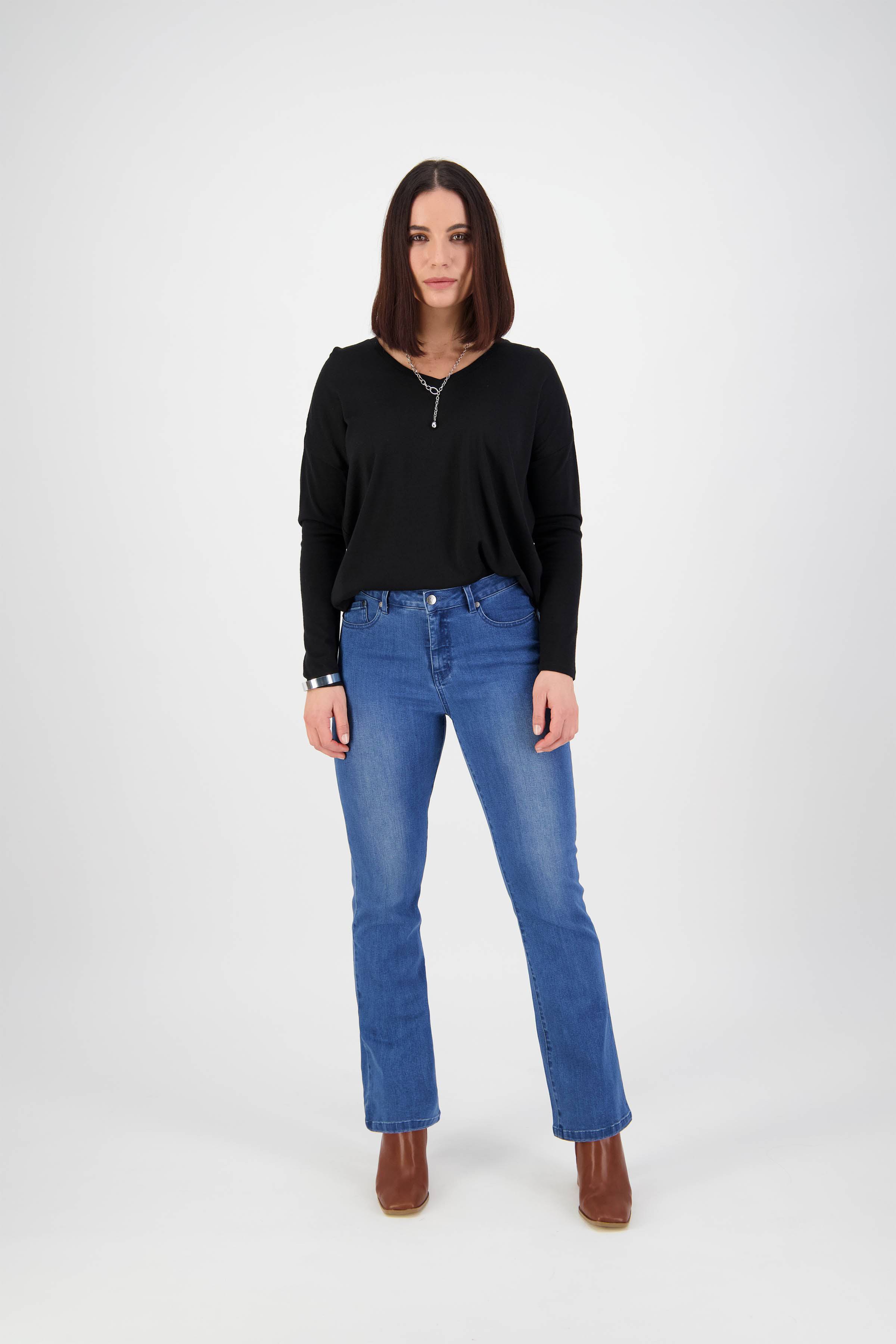 A woman with straight, shoulder-length brown hair stands facing forward in the Vassalli Theory Jean – Blue Denim (With Fade), a black long-sleeve top, brown heeled boots, and a necklace, set against a plain white background.