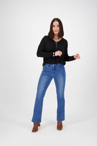A woman with shoulder-length dark hair wears a black long-sleeve top, brown boots, and Vassalli Theory Jean - Blue Denim (With Fade), posing against a plain white background with a slight smile.
