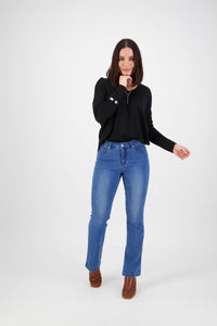 A woman with straight dark hair wears a black long-sleeve top, brown boots, and Vassalli's Theory Jean - Blue Denim (With Fade), standing against a white background and smiling slightly.