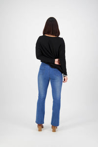 A person with shoulder-length dark hair, in a black long-sleeve top, Vassalli Theory Jean - Blue Denim (With Fade), and tan heeled shoes, stands facing away from the camera against a plain white background.