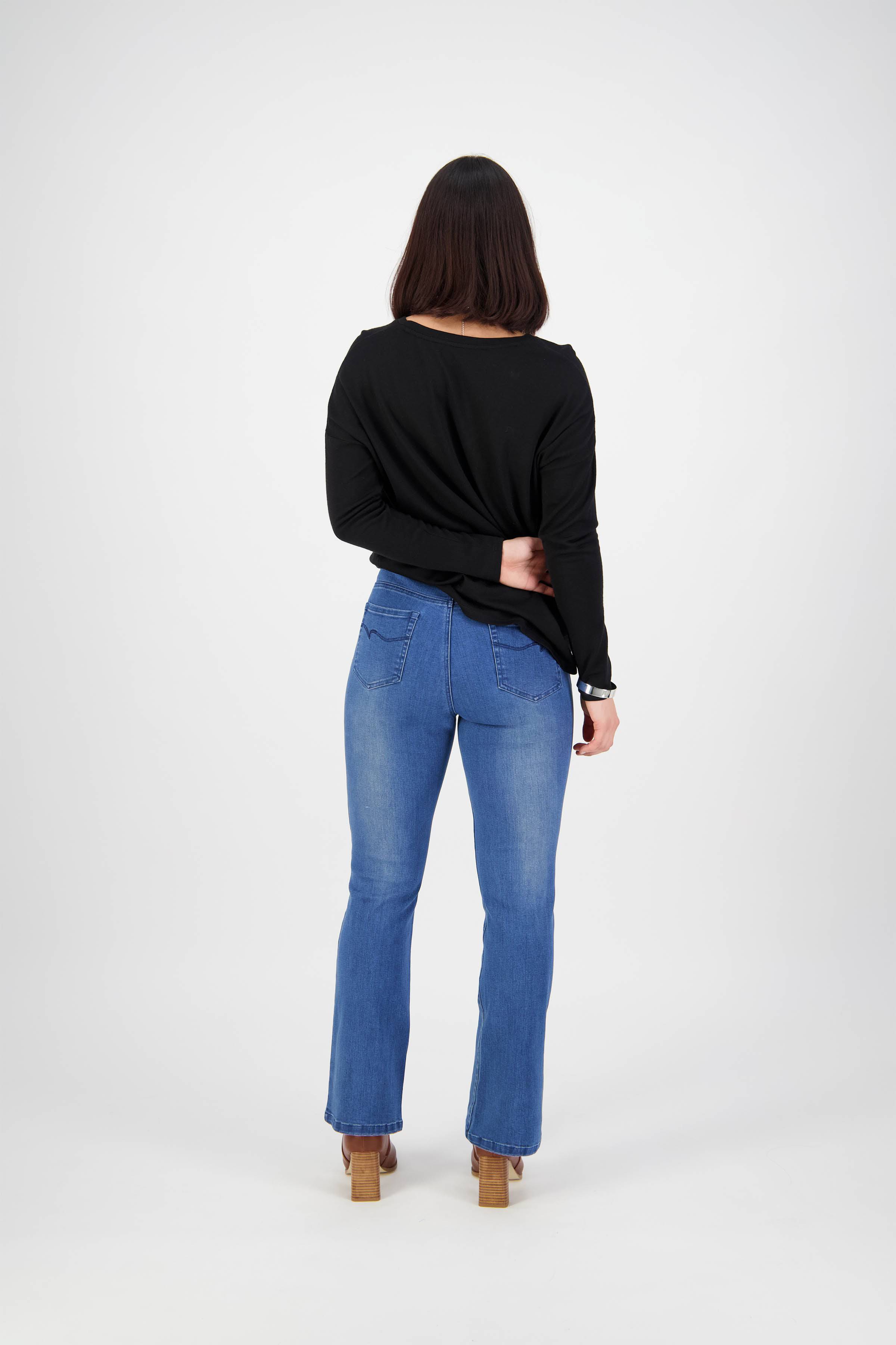 A person with shoulder-length dark hair, in a black long-sleeve top, Vassalli Theory Jean - Blue Denim (With Fade), and tan heeled shoes, stands facing away from the camera against a plain white background.