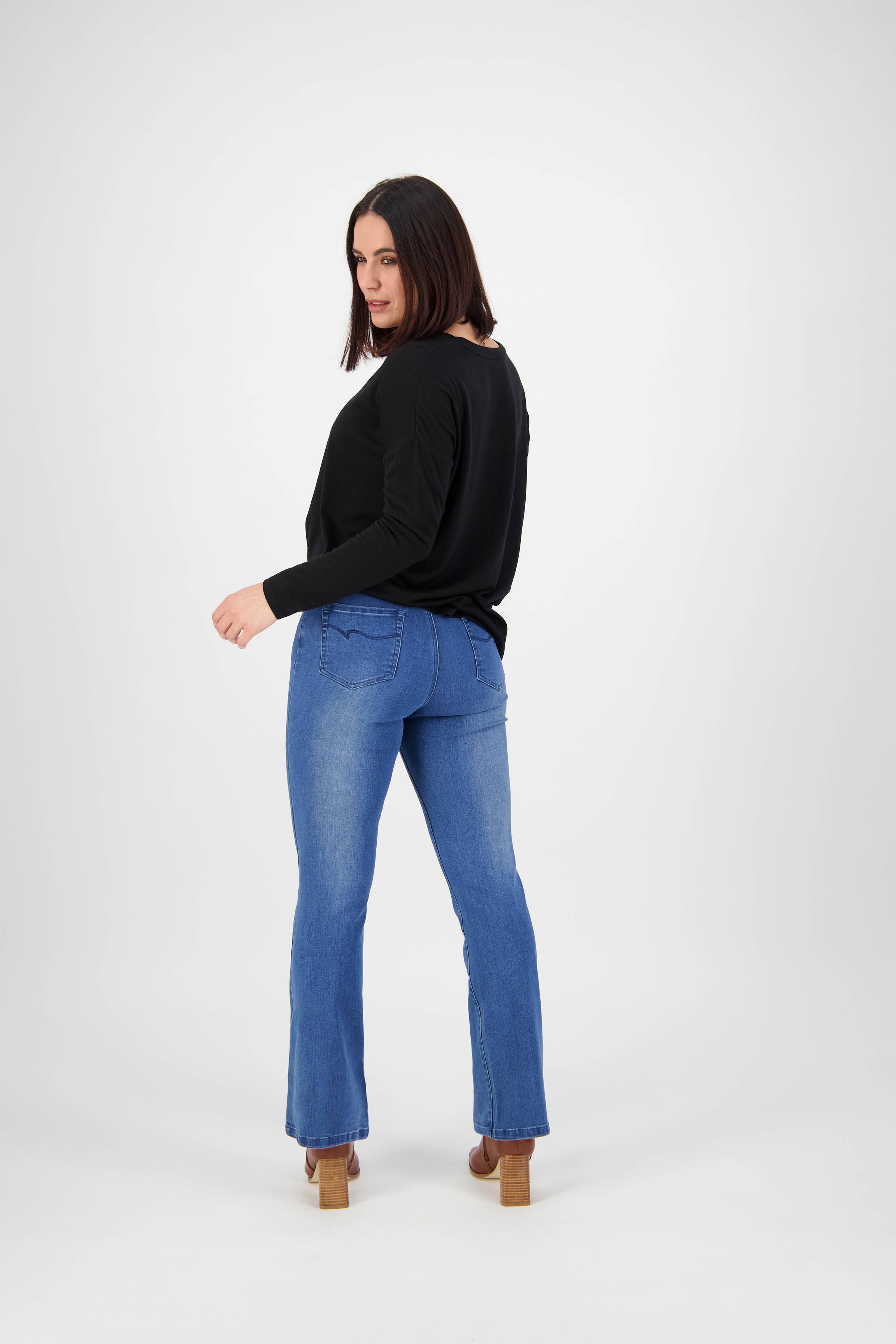 A woman with shoulder-length dark hair stands facing away, glancing over her shoulder. She wears Vassalli's Theory Jean - Blue Denim (With Fade), a black long-sleeve top, and heeled shoes against a plain white background.