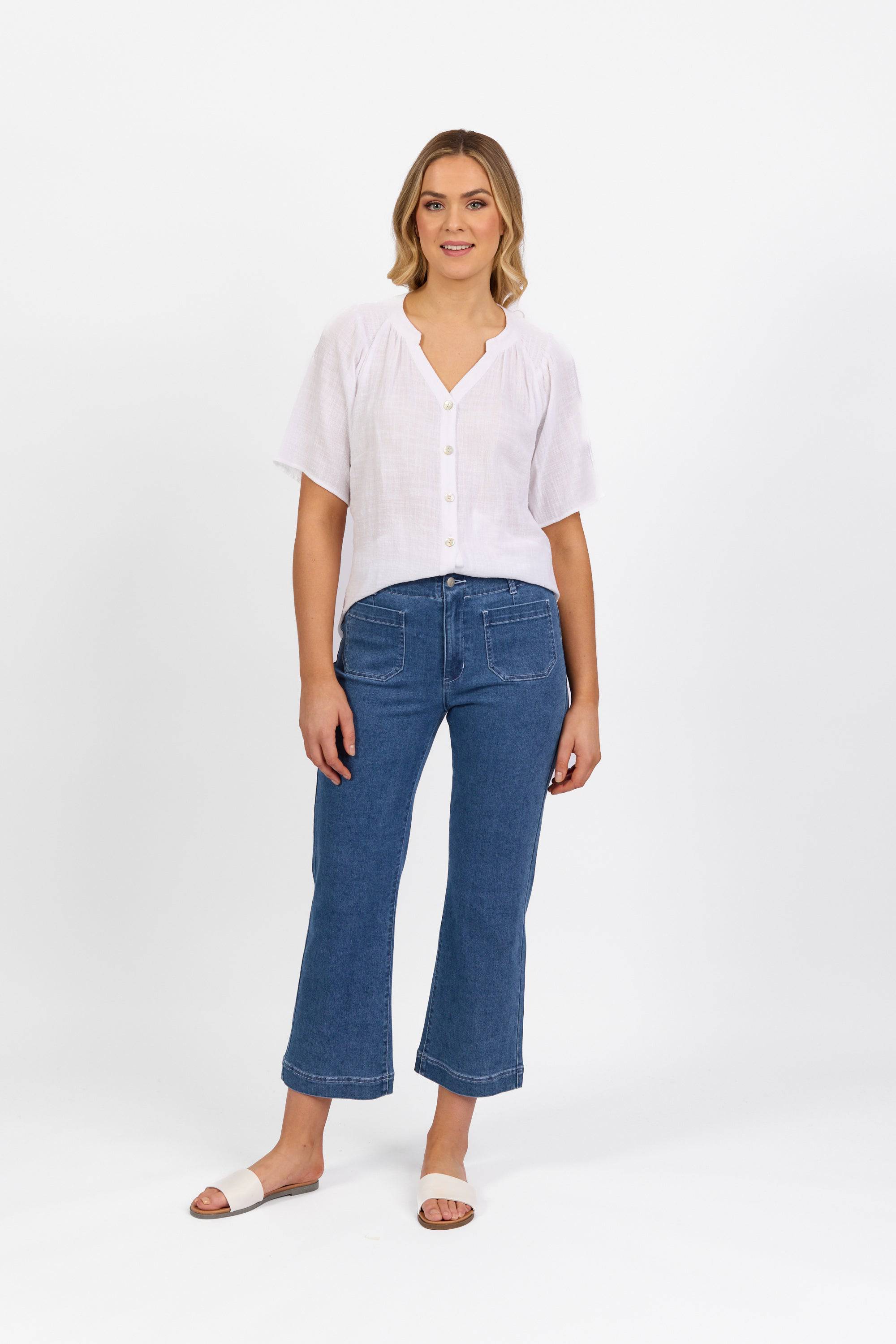 A woman with light brown wavy hair stands against a white background, smiling slightly, wearing Vassalli's Daily Jean in Blue Hue Denim, a white short-sleeve button-up blouse, and white slide sandals.