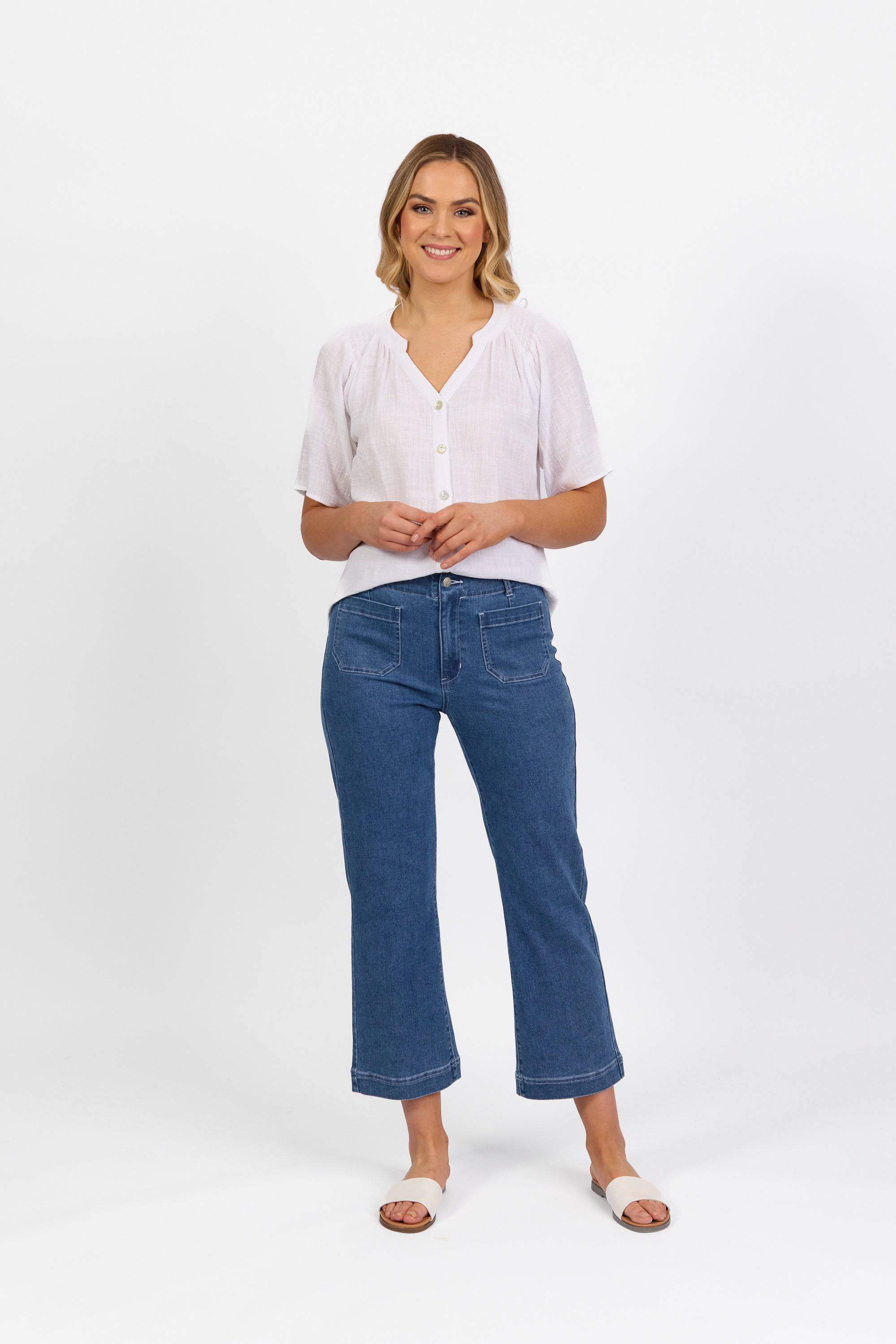A woman with shoulder-length blonde hair smiles against a white background, wearing Vassalli's Daily Jean in Blue Hue Denim, a short-sleeved white blouse, and white sandals. Her hands are loosely clasped in front of her.