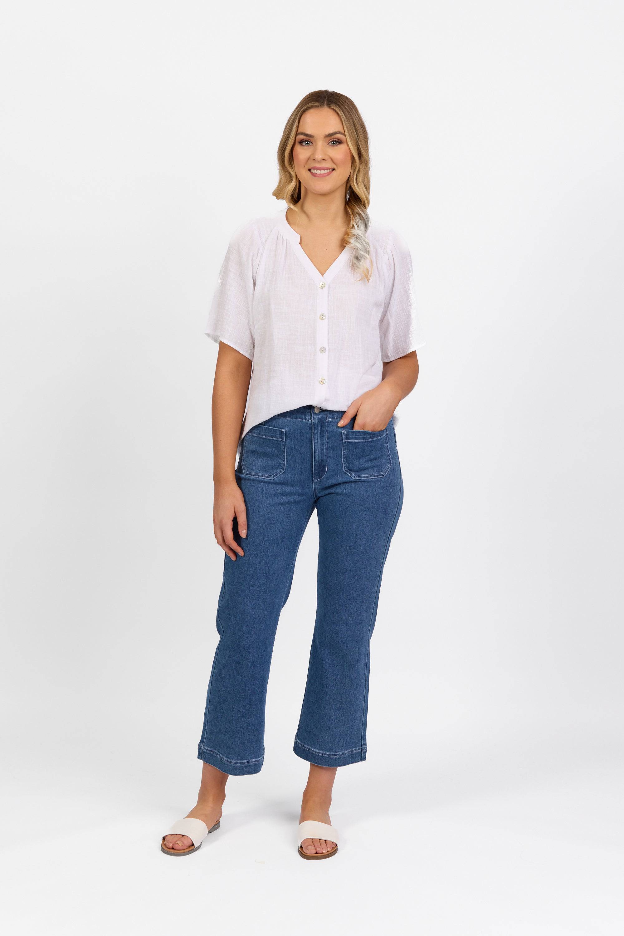 A woman with long blonde hair, wearing a Vassalli Daily Jean - Blue Hue Denim, a white short-sleeve button-up shirt, and white sandals stands smiling against a plain white background.
