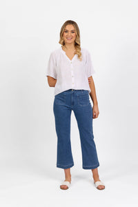 A woman with wavy blonde hair styled to one side smiles against a white background, wearing a white short-sleeve blouse, Vassalli Daily Jean - Blue Hue Denim straight leg jeans, and white sandals.