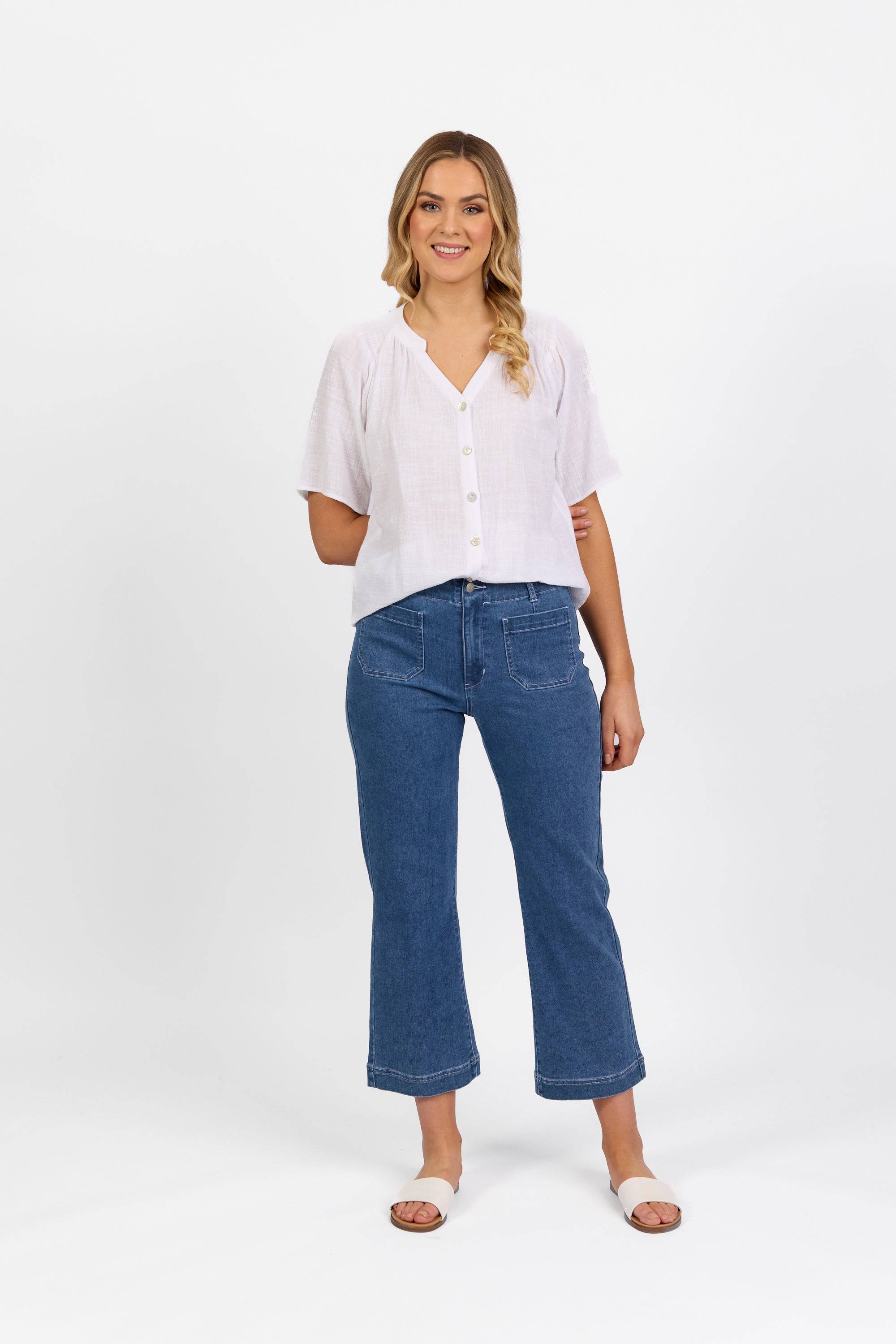 A woman with wavy blonde hair styled to one side smiles against a white background, wearing a white short-sleeve blouse, Vassalli Daily Jean - Blue Hue Denim straight leg jeans, and white sandals.