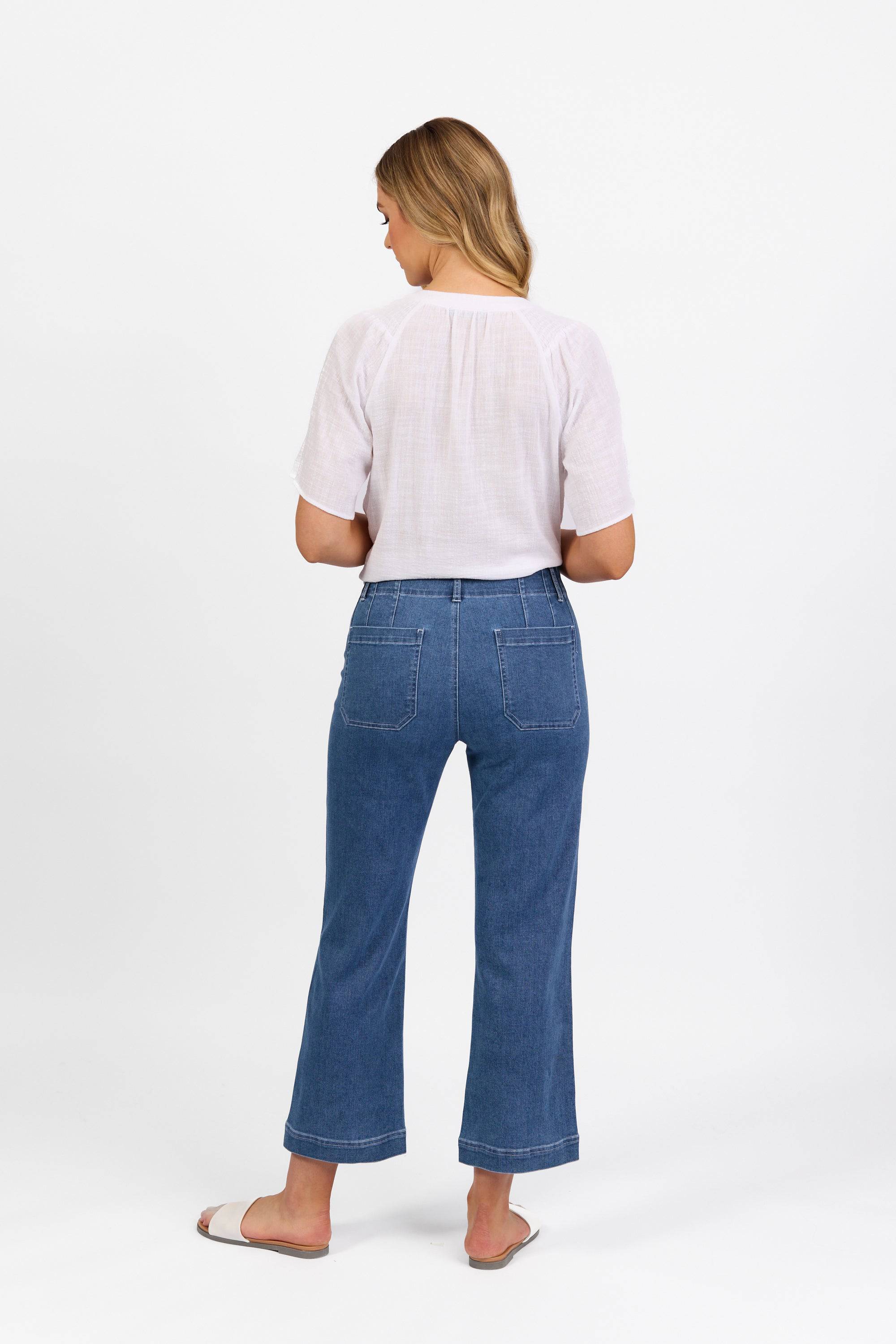 A woman with long blonde hair stands facing away, wearing a white short-sleeve blouse, tan sandals, and Vassalli Daily Jean - Blue Hue Denim against a plain white background.