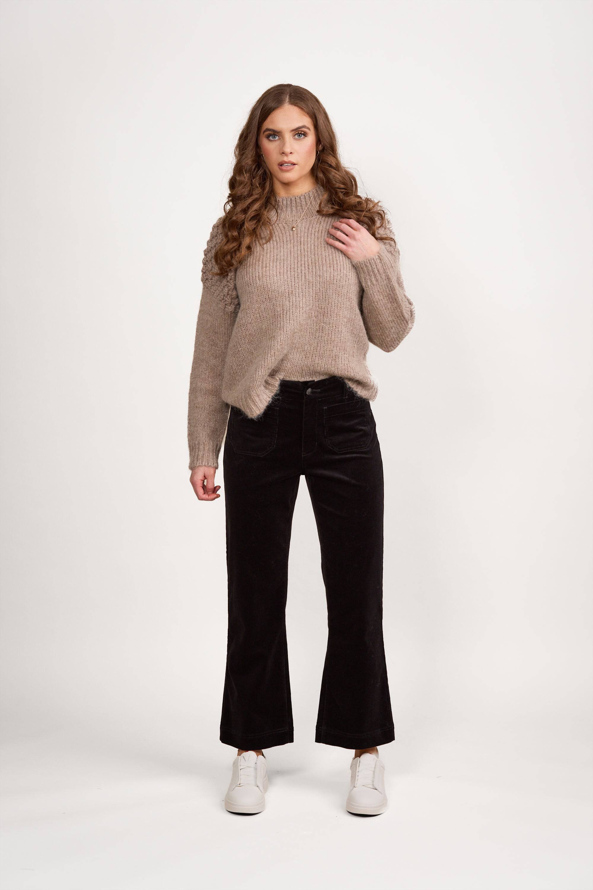 A woman with long curly hair wears a beige knit sweater, Vassalli Daily Cord Pant in black, and white sneakers, standing against a plain white background.