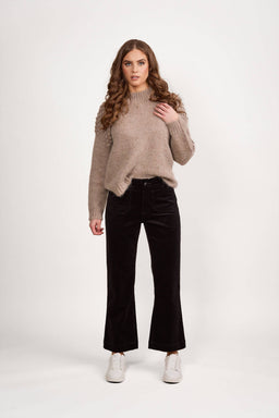 A woman with long curly hair wears a beige knit sweater, Vassalli Daily Cord Pant in black, and white sneakers, standing against a plain white background.