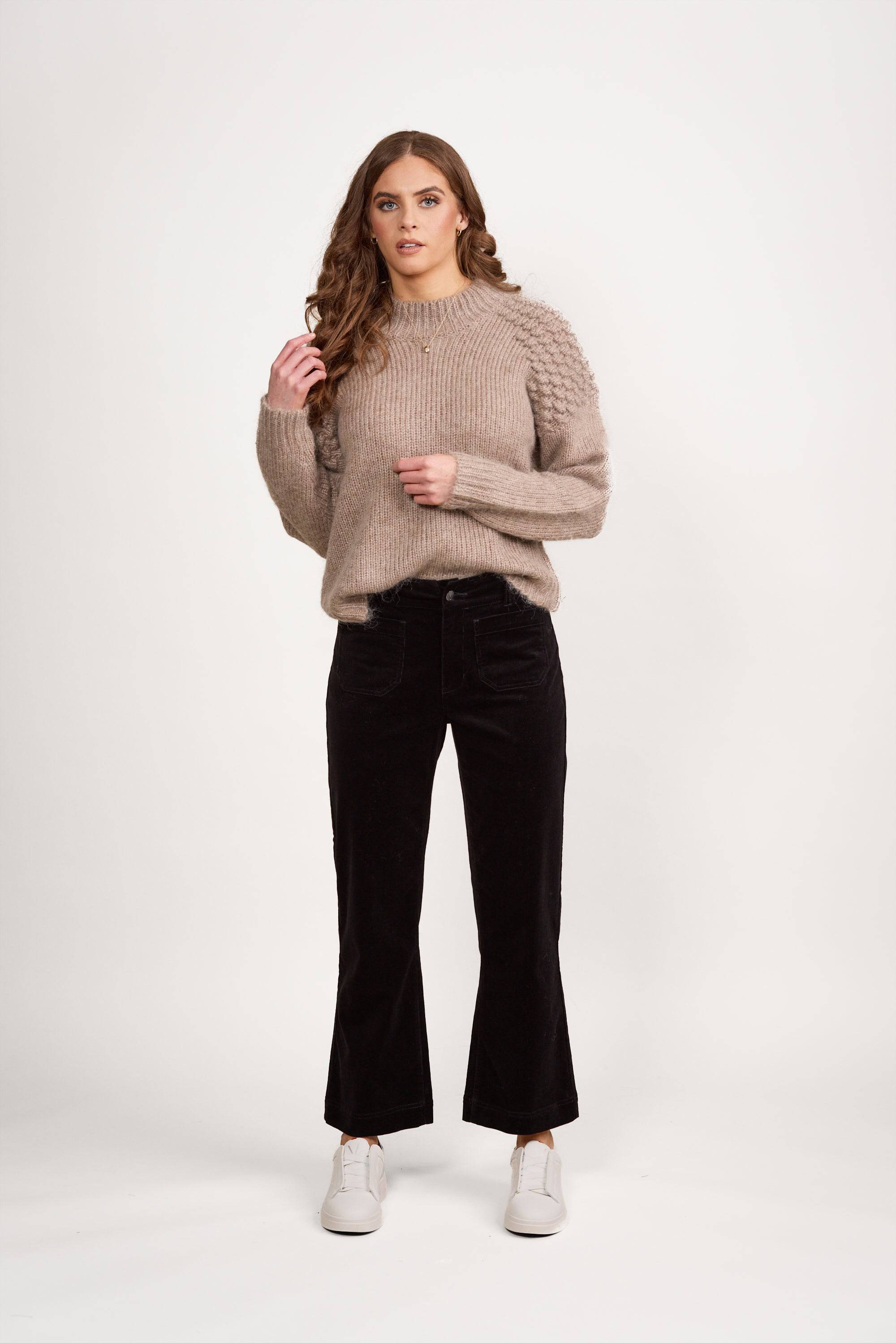 A woman with long wavy brown hair wears a beige knit sweater and Vassalli Daily Cord Pant - Black. She pairs them with white shoes and stands against a plain white background, looking forward with one hand near her hair.