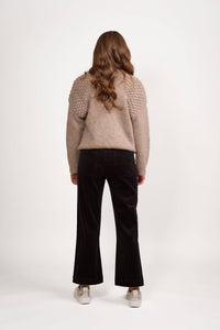 A person with long wavy brown hair wears a textured beige sweater and Vassalli's Daily Cord Pant in Black, featuring straight legs and front patch pockets, plus light sneakers, standing back to camera against a white background.