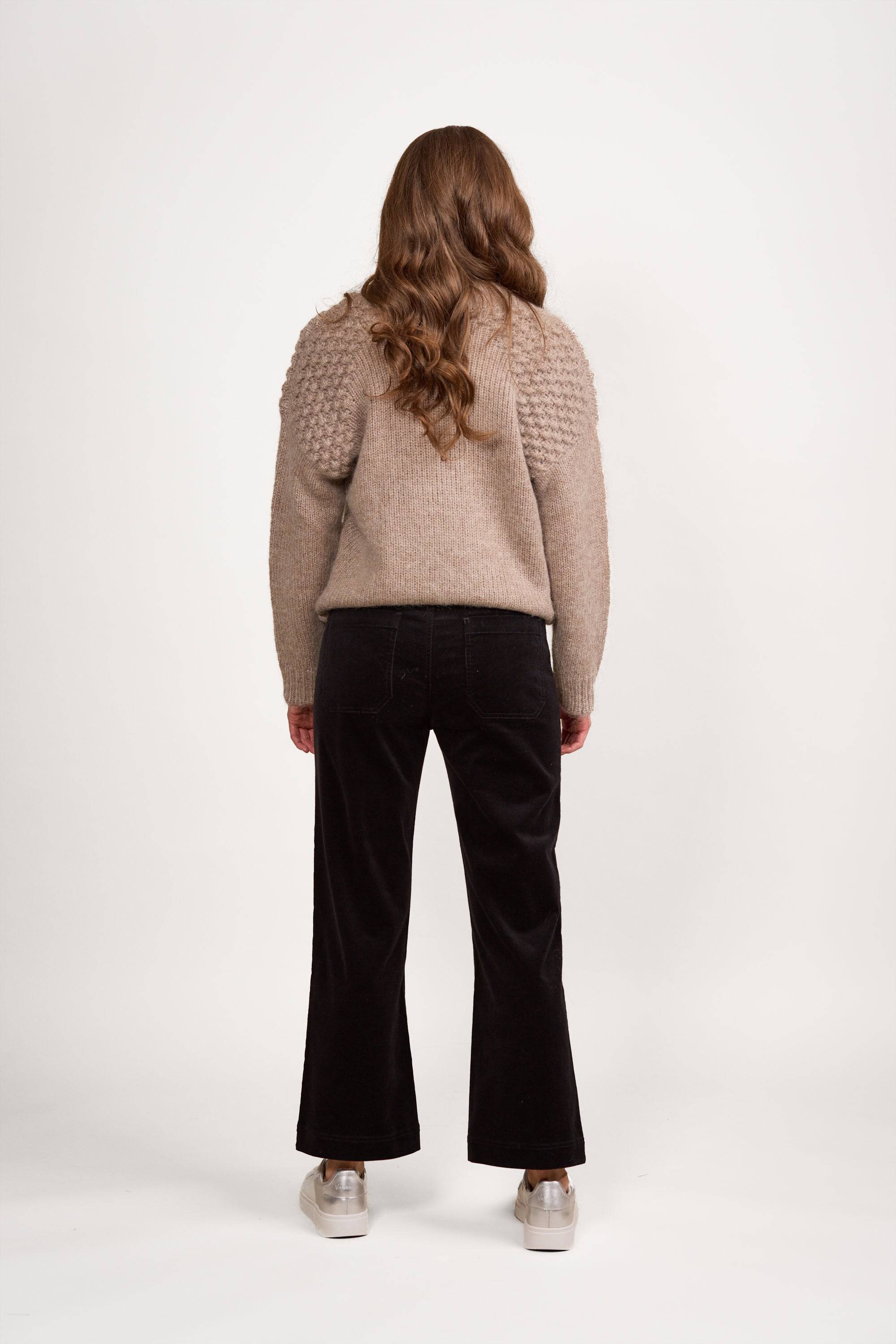 A person with long wavy brown hair wears a textured beige sweater and Vassalli's Daily Cord Pant in Black, featuring straight legs and front patch pockets, plus light sneakers, standing back to camera against a white background.