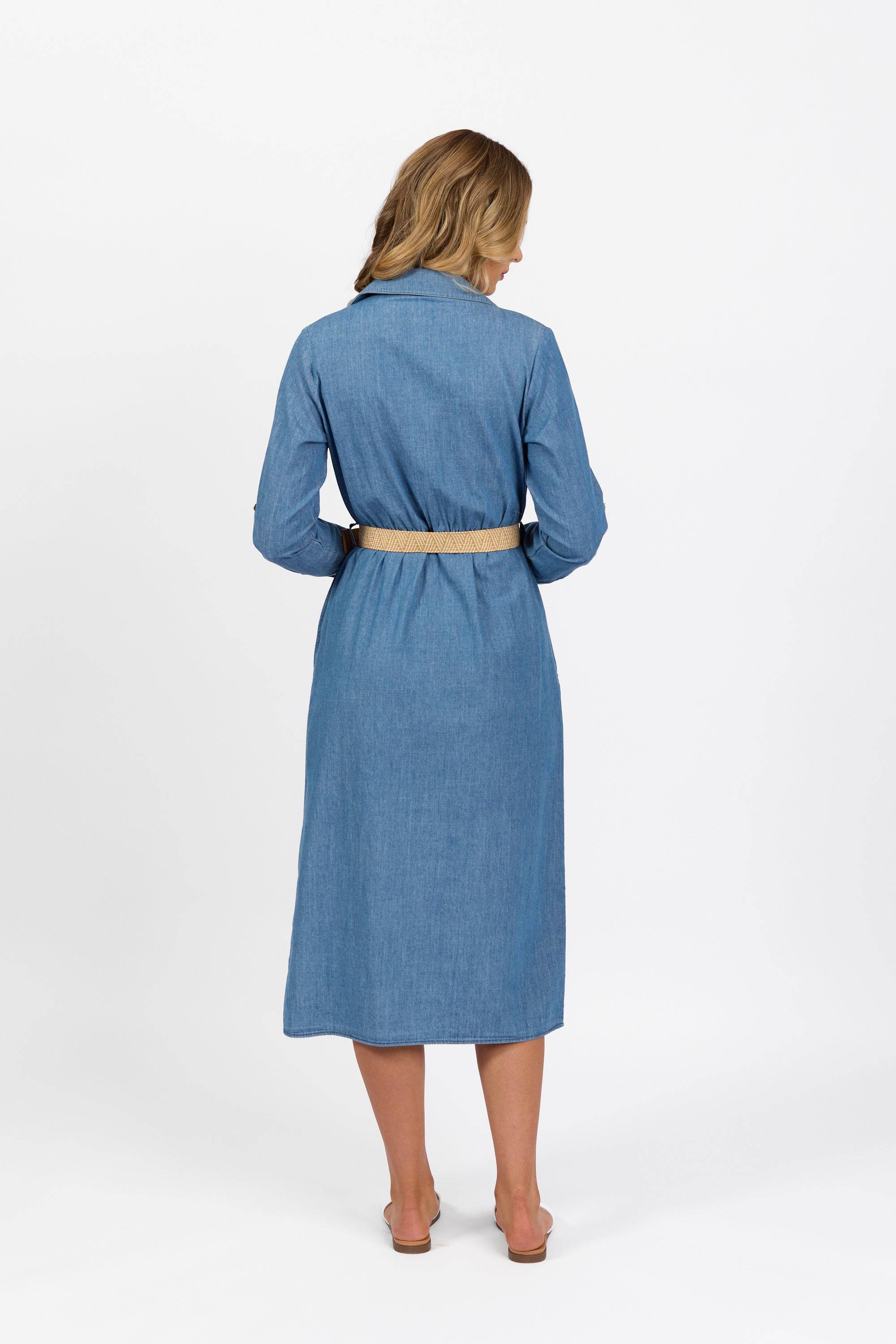 A woman with shoulder-length blonde hair stands barefoot on a white background, wearing the Vassalli Long Sleeve Dress - Blue Wash with a beige belt.