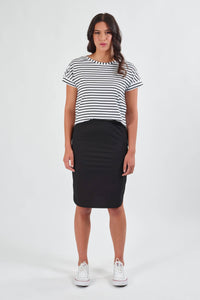 A woman faces forward against a plain white background, wearing the Vassalli Poplin Comfort Skirt in black, a black-and-white striped short-sleeve shirt, and white sneakers.